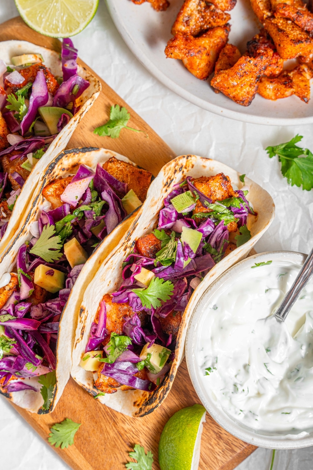 A wooden server with three mahi mahi tacos next to a bowl of sour cream sauce. Everything is garnished with cilantro and there is an extra plate of fried mahi mahi on the side.