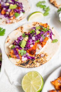 An open mahi mahi taco on a piece of parchment paper on a marble counter. There is an additional taco and lime wedges on the side.