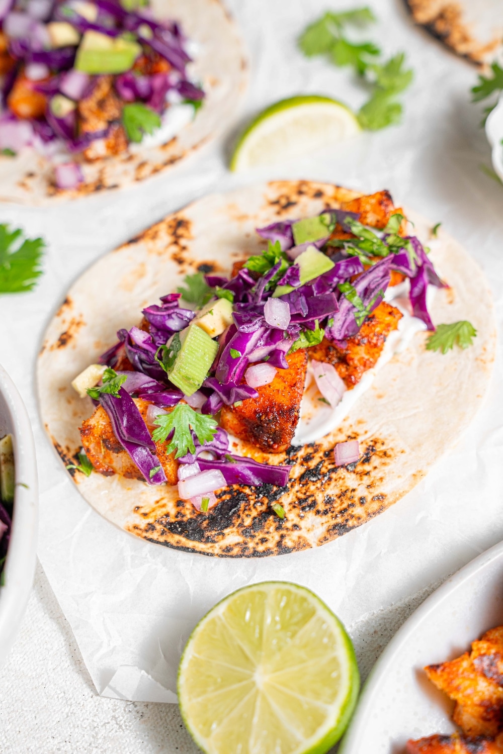 An open mahi mahi taco on a piece of parchment paper on a marble counter. There is an additional taco and lime wedges on the side.