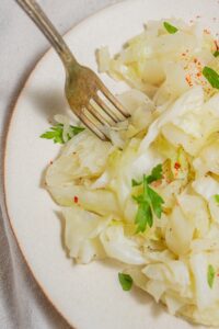 A close up of a plate of steamed cabbage on a white plate with a fork digging into the cabbage. The cabbage is garnished with fresh parsley.