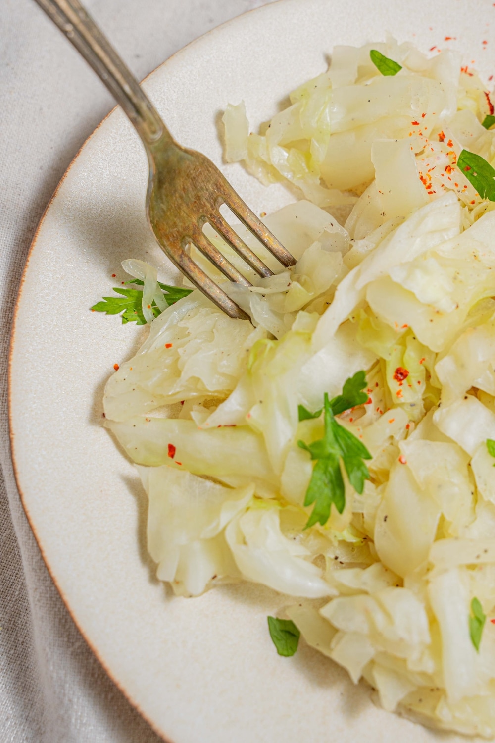A close up of a plate of steamed cabbage on a white plate with a fork digging into the cabbage. The cabbage is garnished with fresh parsley.