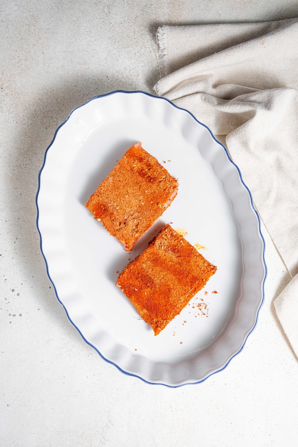 An overhead shot of a baking dish with two raw seasoned mahi mahi fillets. The baking dish is on a marble counter with a cloth napkin next to it.