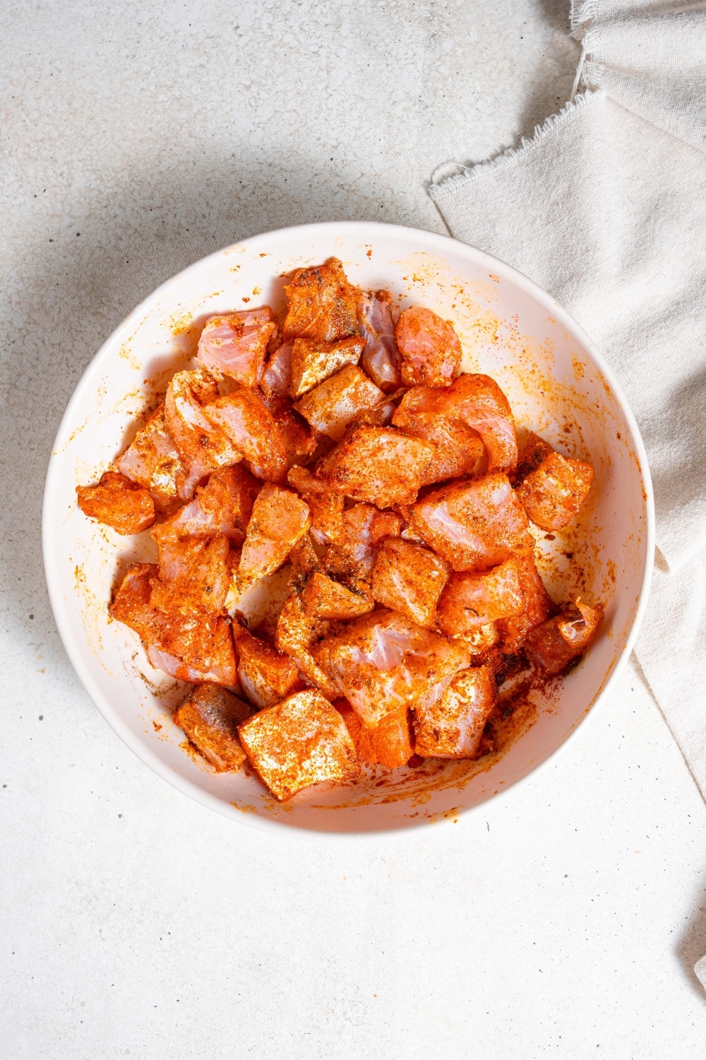 An overhead shot of cubed raw mahi mahi marinating in a seasoning mix in a ceramic bowl on a marble counter.