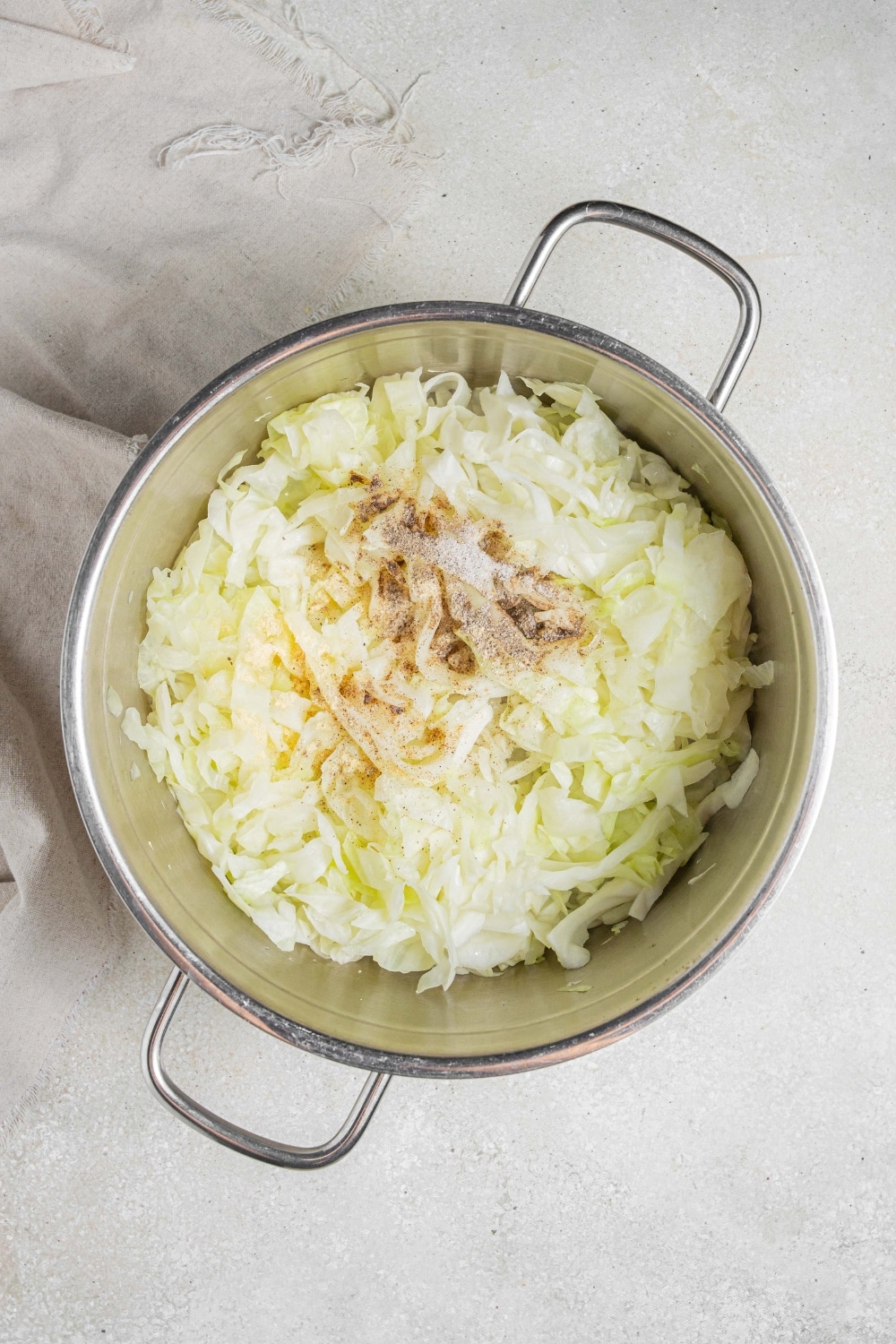 An overhead shot of a large pot filled with steamed cabbage topped with seasonings. The pot is on a marble counter with a cloth napkin off to the side.