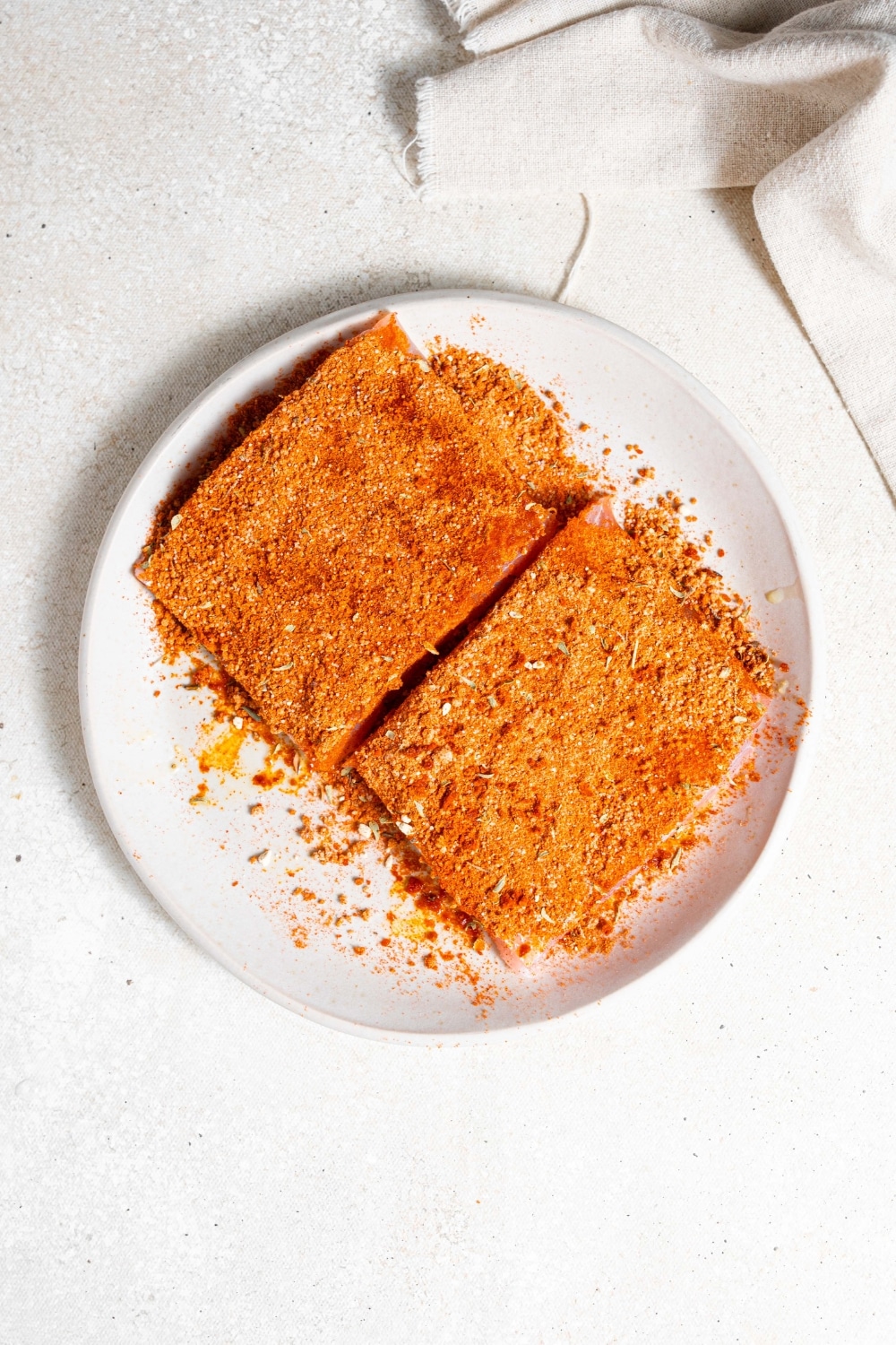 An overhead shot of a ceramic plate with two mahi mahi filets seasoned with cajun spices. The plate is on a marble counter with a cloth napkin next to it.
