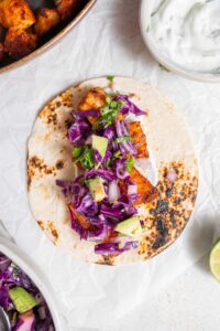 An open mahi mahi taco topped with avocado slaw. The taco is on a piece of parchment paper on a marble counter with a bowl of sour cream sauce on the side.