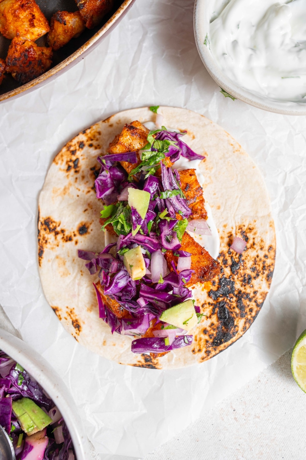 An open mahi mahi taco topped with avocado slaw. The taco is on a piece of parchment paper on a marble counter with a bowl of sour cream sauce on the side.