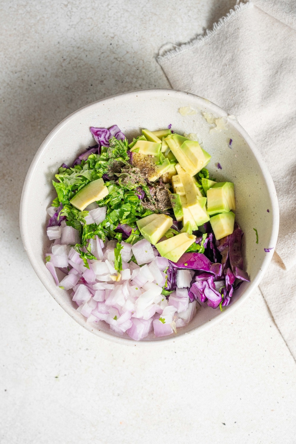 An overhead shot of bowl filled with ingredients to make avocado slaw on a marble counter with a cloth napkin off to the side.
