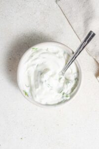 An overhead shot of a bowl of sour cream sauce with a spoon dipped in it. The bowl is on a marble counter with a cloth napkin off to the side.