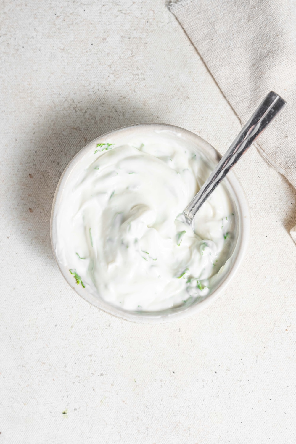 An overhead shot of a bowl of sour cream sauce with a spoon dipped in it. The bowl is on a marble counter with a cloth napkin off to the side.