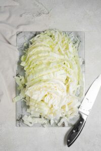 An overhead shot of a marble cutting board on a marble counter. The cutting board has chopped cabbage with a knife to the side. There is a cloth napkin next to the cutting board.