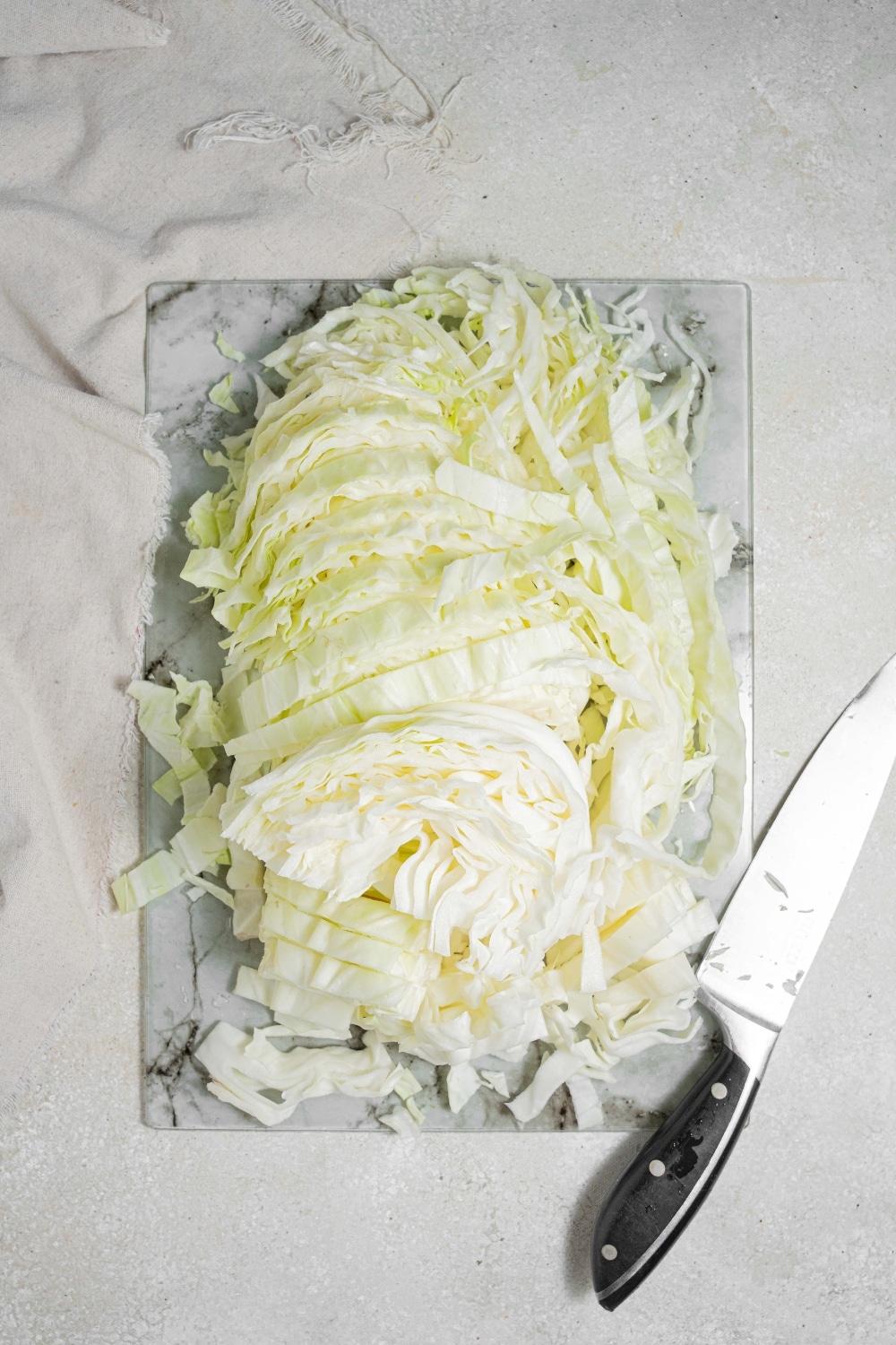 An overhead shot of a marble cutting board on a marble counter. The cutting board has chopped cabbage with a knife to the side. There is a cloth napkin next to the cutting board.