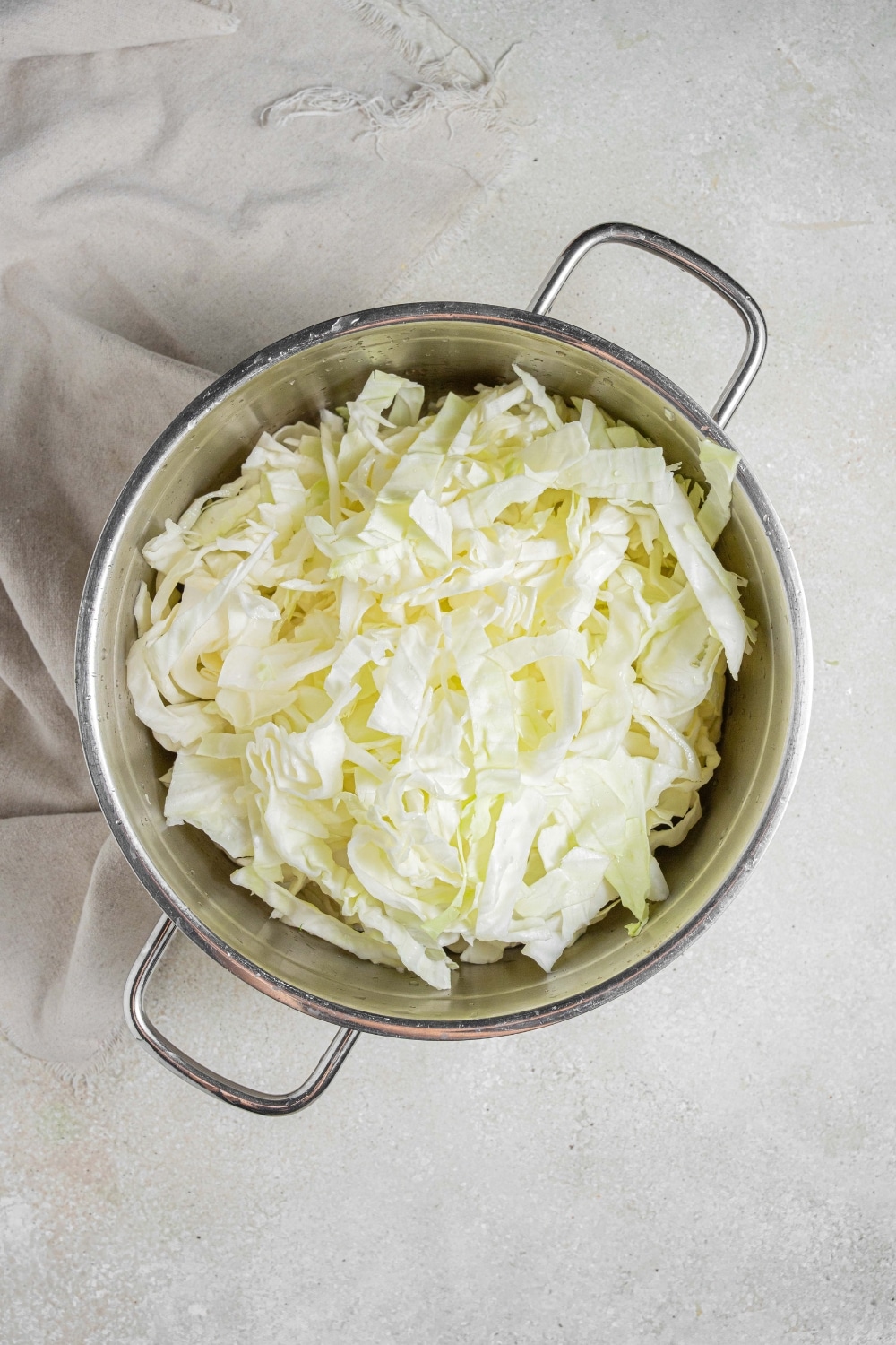 An overhead shot of a large pot filled with raw sliced cabbage. The pot is on a marble counter with a cloth napkin off to the side.
