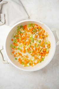An overhead shot of a large stock pot with chopped carrots, onions, and celery in olive oil on a marble counter with a cloth napkin on the side.
