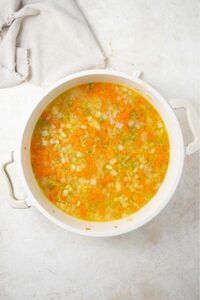 An overhead shot of a large stock pot filled with chicken stock and chopped celery, onions, and carrots on a marble counter with a cloth napkin on the side.