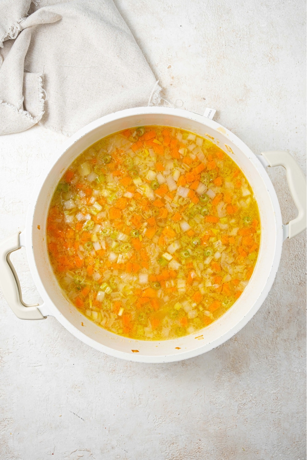 An overhead shot of a large stock pot filled with chicken stock and chopped celery, onions, and carrots on a marble counter with a cloth napkin on the side.