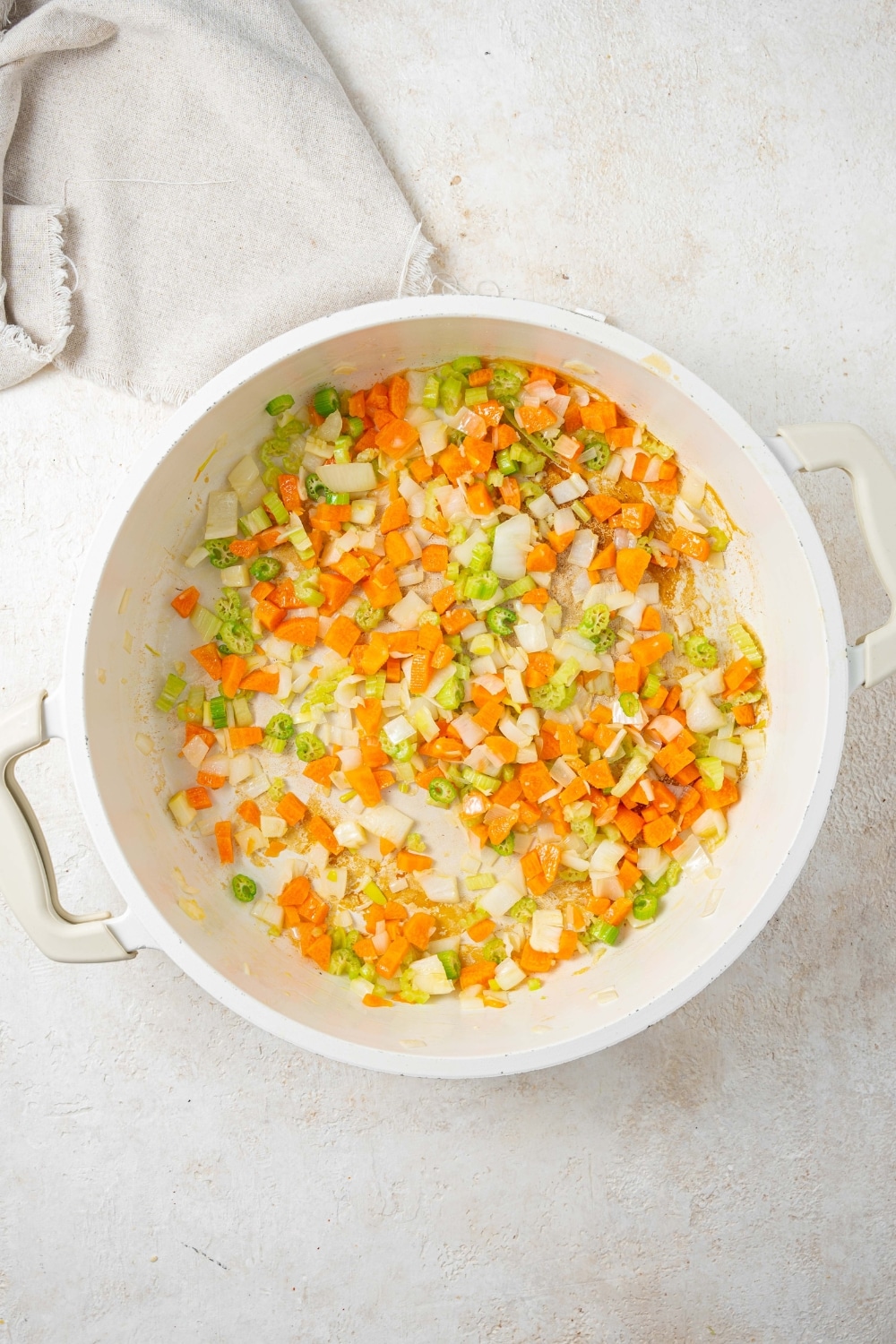 An overhead shot of a large stock pot with chopped carrots, onions, and celery in olive oil on a marble counter with a cloth napkin on the side.