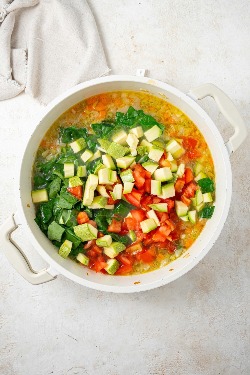 An overhead shot of a large stock pot with chopped veggies over a chicken stock mixture on a marble counter with a cloth napkin on the side.