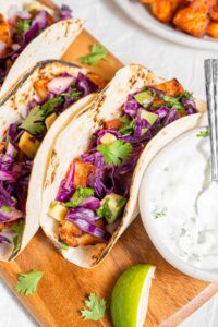 A wooden server with three mahi mahi tacos next to a bowl of sour cream sauce. Everything is garnished with cilantro and there is an extra plate of fried mahi mahi on the side.