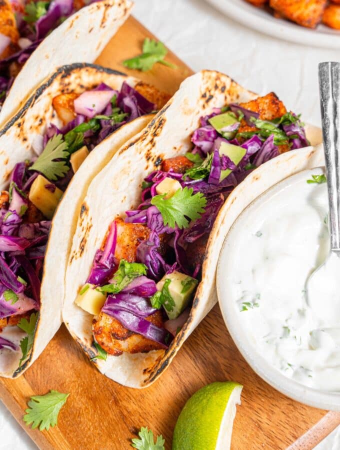A wooden server with three mahi mahi tacos next to a bowl of sour cream sauce. Everything is garnished with cilantro and there is an extra plate of fried mahi mahi on the side.