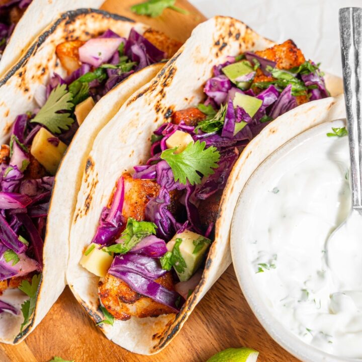 A wooden server with three mahi mahi tacos next to a bowl of sour cream sauce. Everything is garnished with cilantro and there is an extra plate of fried mahi mahi on the side.