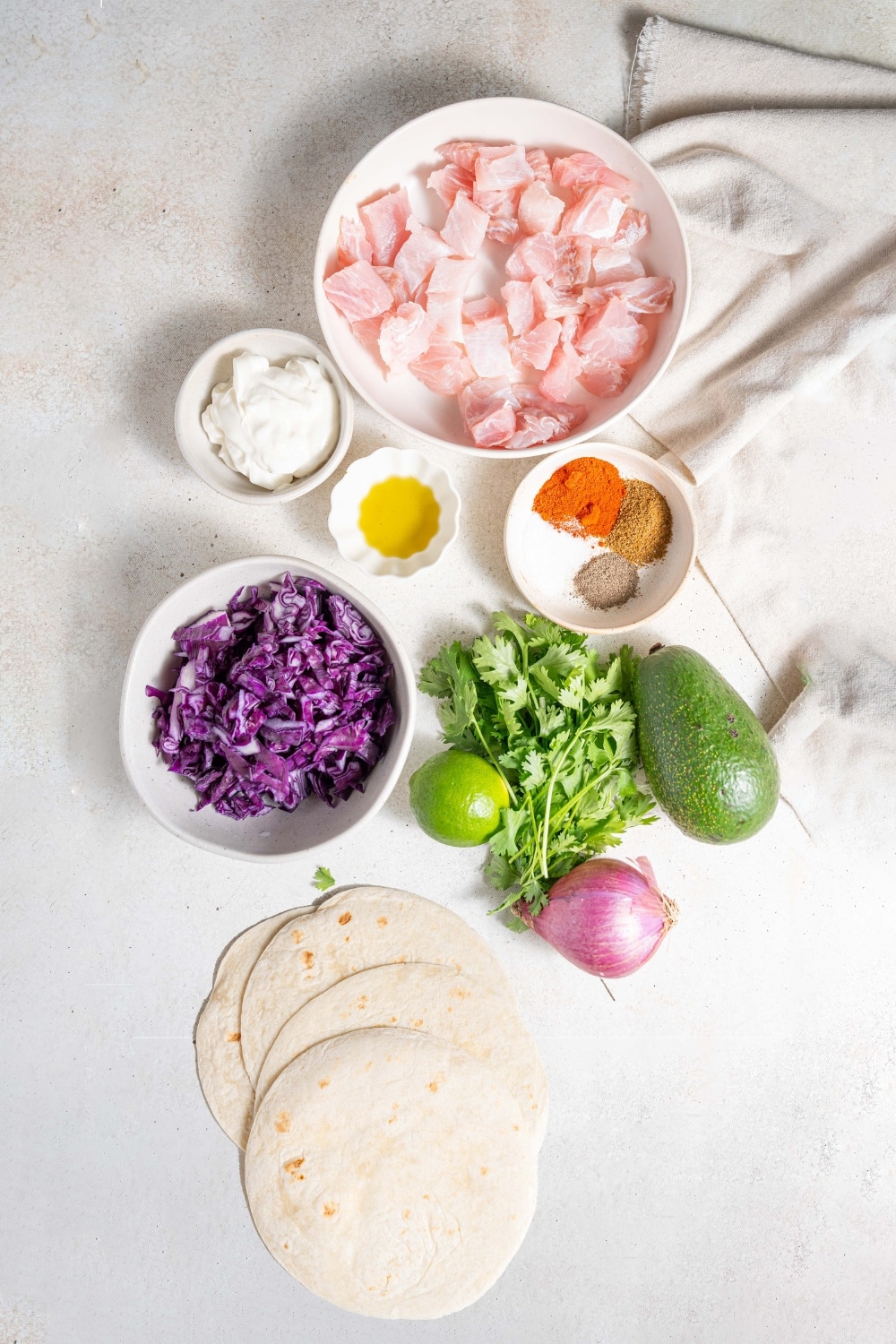 An overhead shot of several bowls containing various ingredients to make mahi mahi tacos including raw cubed mahi mahi, sour cream, shredded purple cabbage, and seasonings.