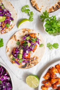 An open mahi mahi fish taco topped with avocado slaw on a piece of parchment paper on a marble counter. There is an additional taco and plate of cooked mahi mahi on the side with cilantro sprinkled over everything.