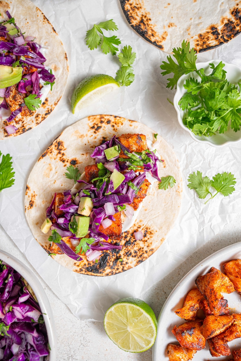 An open mahi mahi fish taco topped with avocado slaw on a piece of parchment paper on a marble counter. There is an additional taco and plate of cooked mahi mahi on the side with cilantro sprinkled over everything.