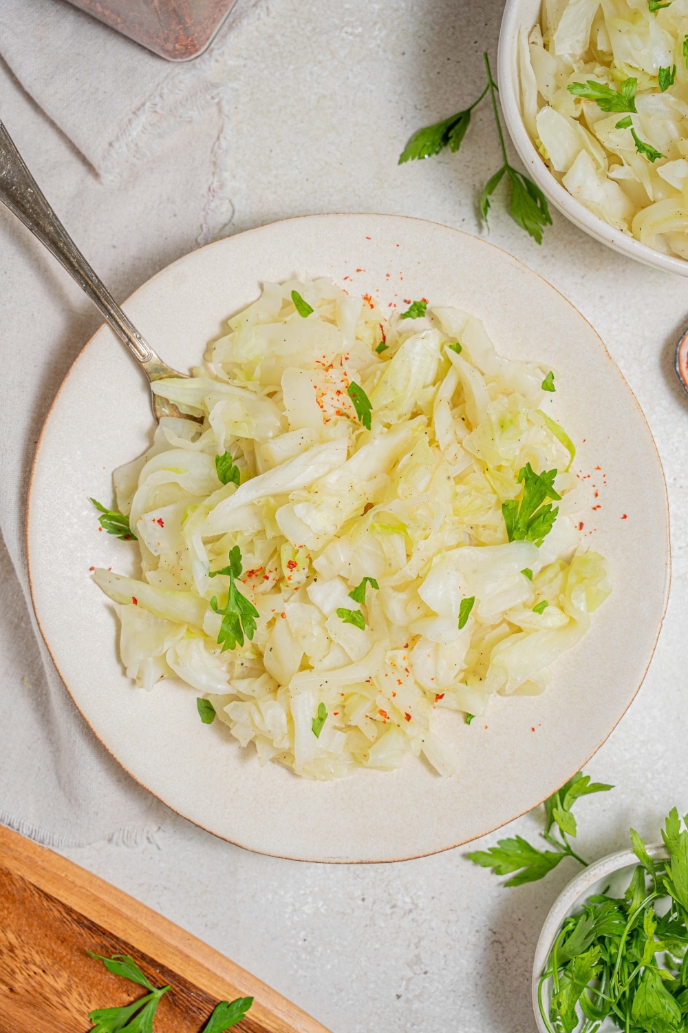 An overhead shot of a white plate with steamed cabbage on a marble counter. There is a fork digging into the cabbage. There is a cloth napkin under the plate with an additional bowl of steamed cabbage and another bowl of fresh parsley off to the side.