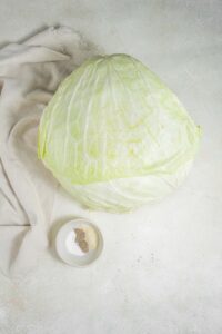An overhead shot of a head of cabbage next to a small bowl of seasonings on a marble counter with a cloth napkin to the side.