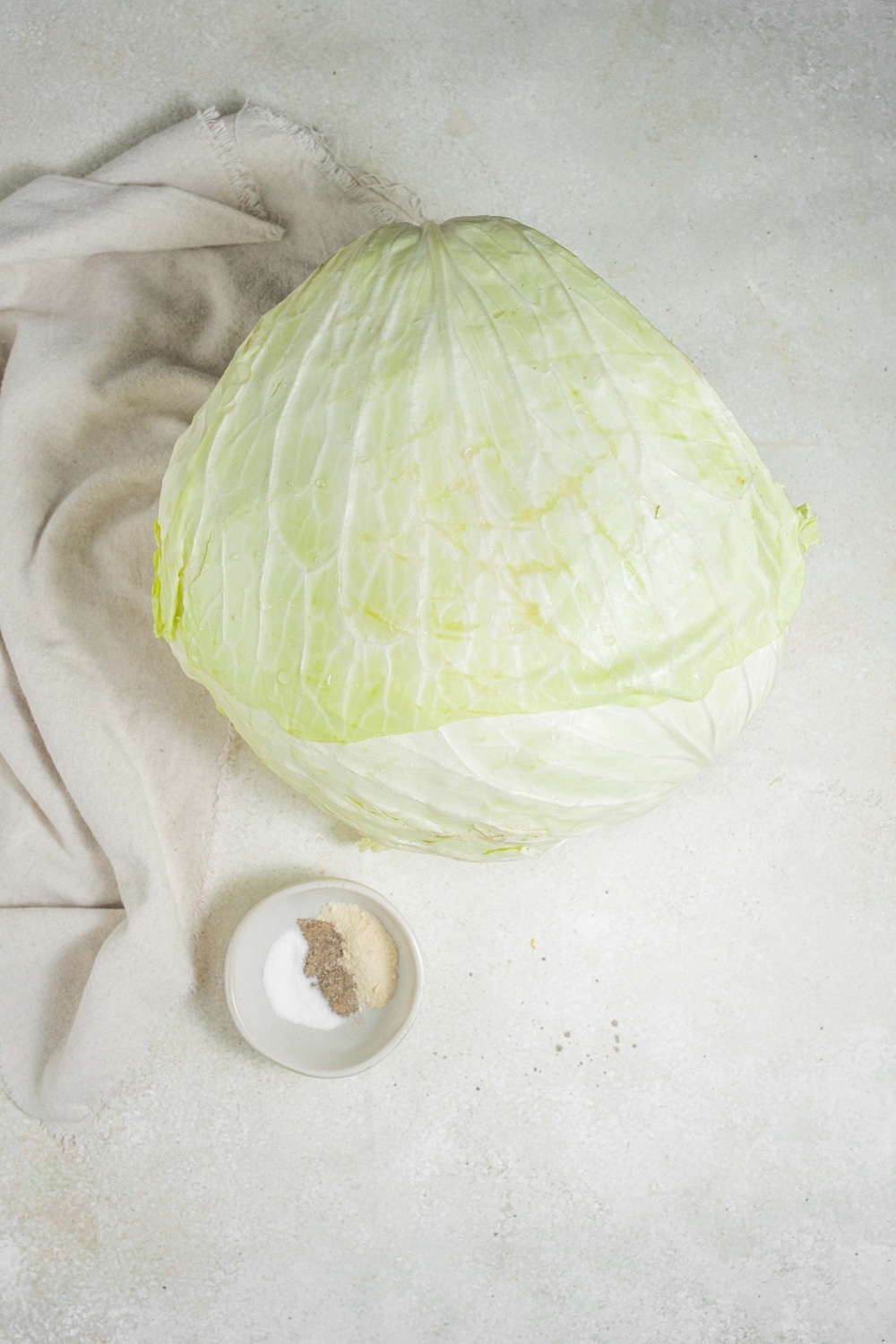 An overhead shot of a head of cabbage next to a small bowl of seasonings on a marble counter with a cloth napkin to the side.