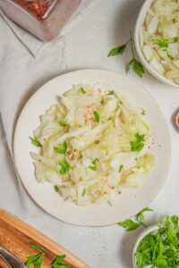 An overhead shot of a white plate with steamed cabbage on a marble counter. There is a cloth napkin under the plate with an additional bowl of steamed cabbage and another bowl of fresh parsley off to the side.