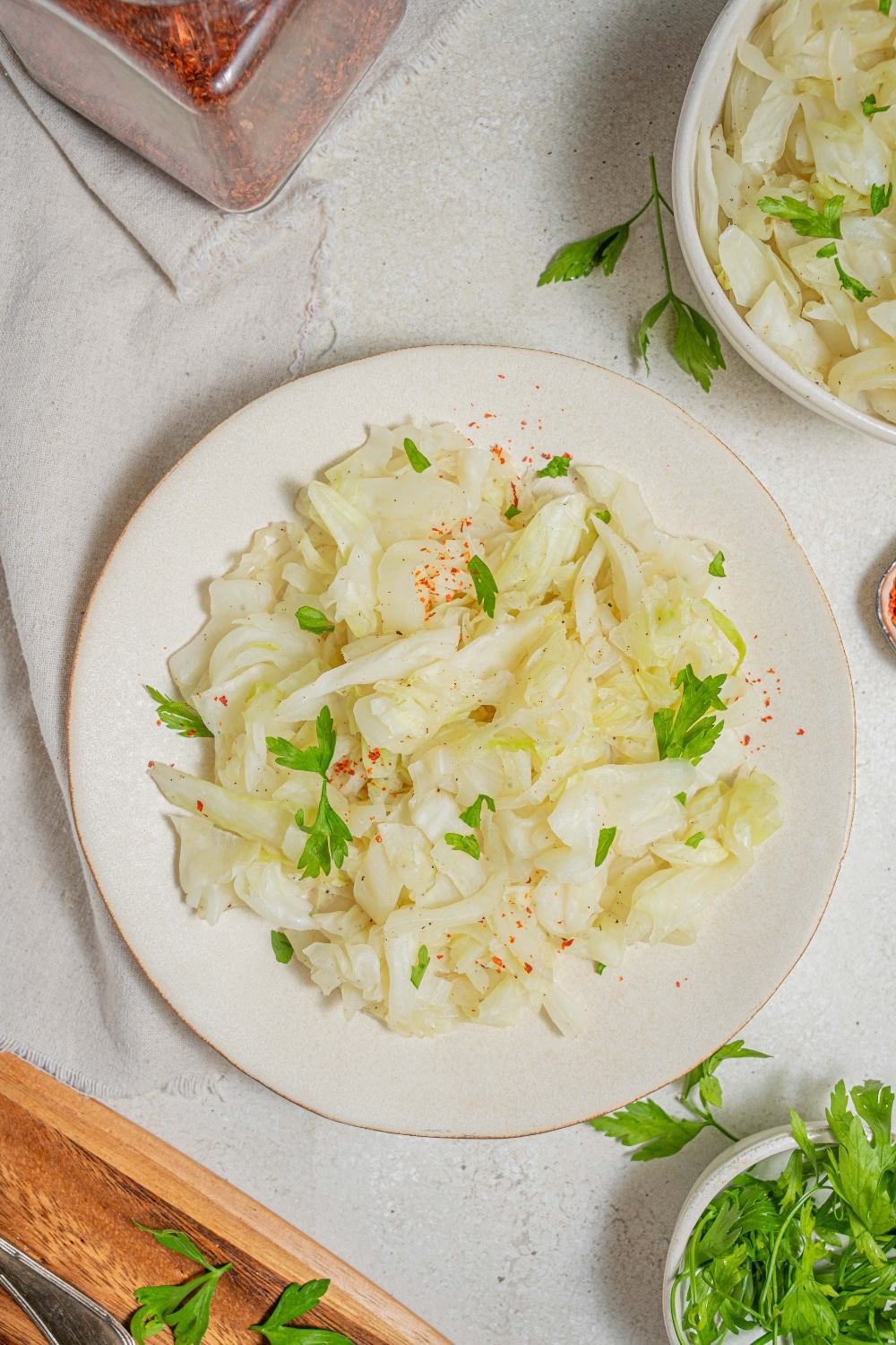 An overhead shot of a white plate with steamed cabbage on a marble counter. There is a cloth napkin under the plate with an additional bowl of steamed cabbage and another bowl of fresh parsley off to the side.