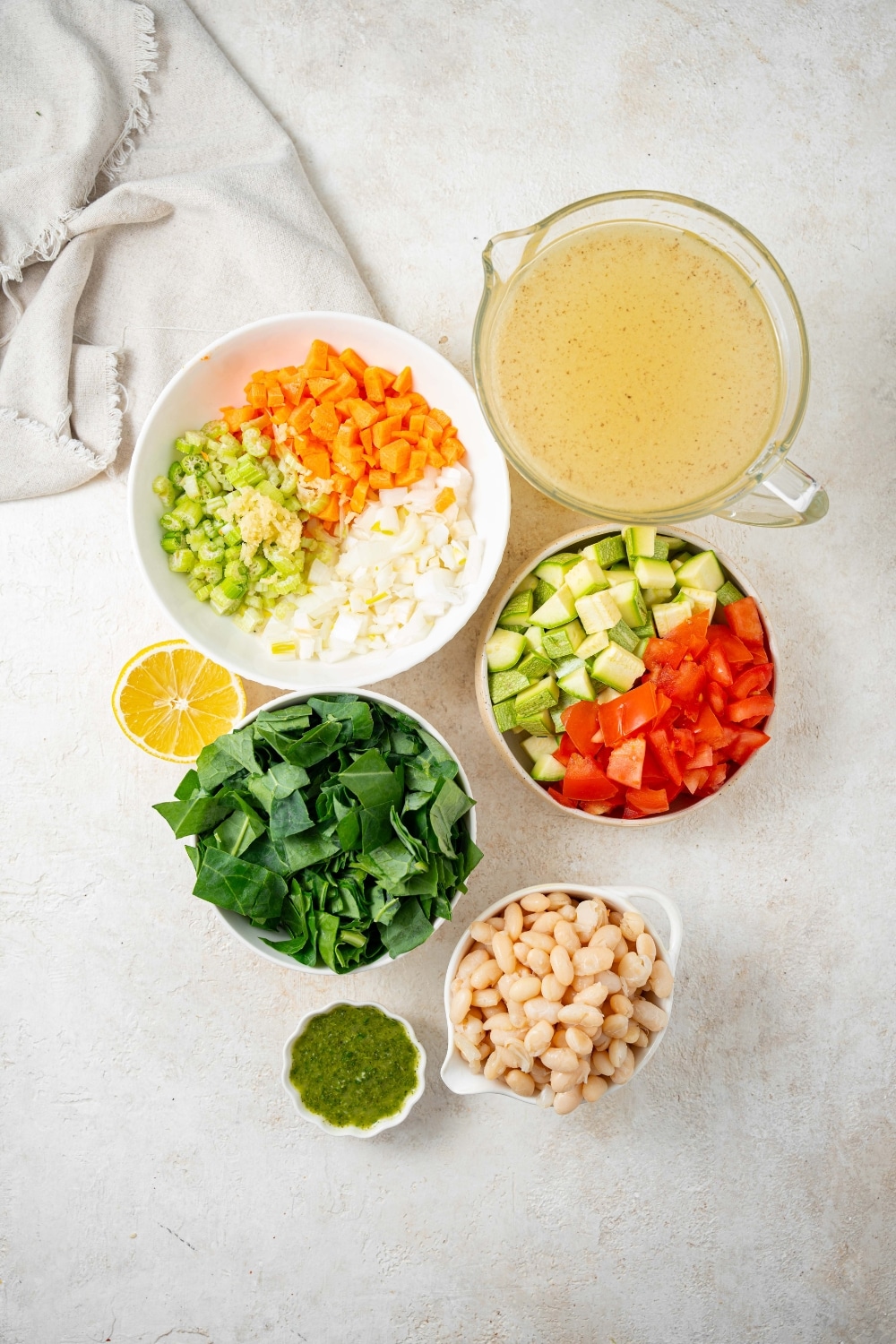 An overhead shot of several bowls of various sizes containing ingredients for vegetable weight loss soup including chicken stock, chopped celery, carrot, onion, red pepper, and zucchini, and white beans.