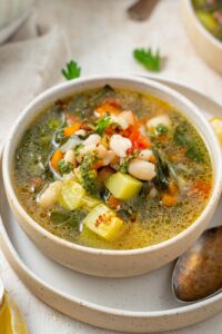 A bowl of vegetable weight loss soup in a ceramic bowl on a ceramic plate. There is a spoon on the plate with a cloth napkin on the side.