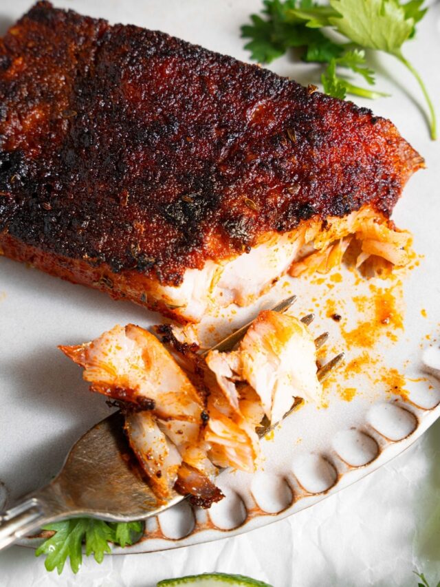 A close up of blackened mahi mahi on a ceramic plate with a fork taking a bite of fish. The plate is garnished with parsley and lime.