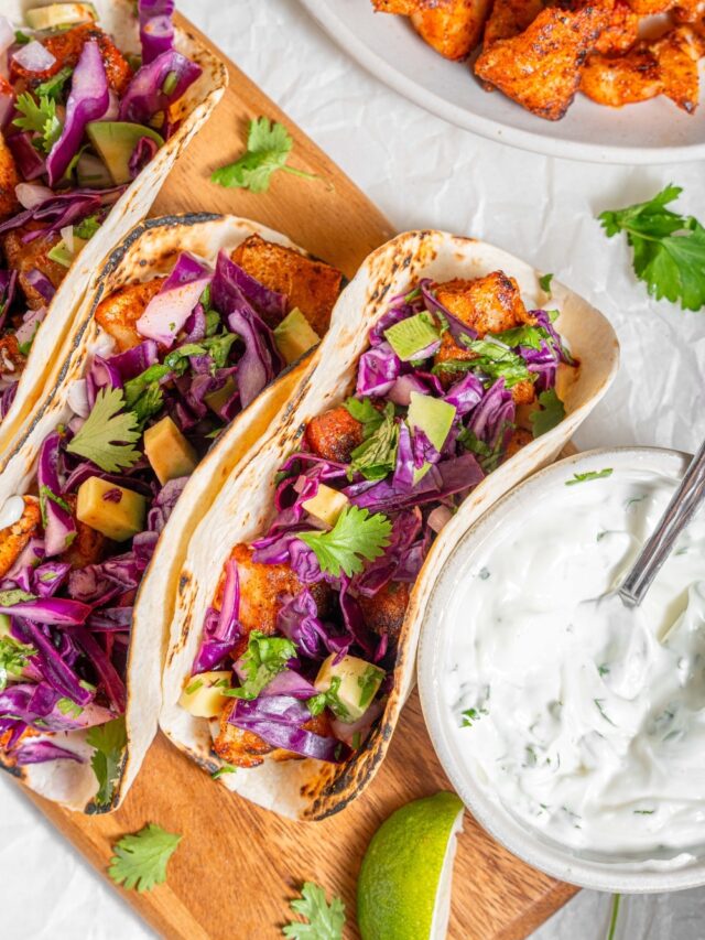 A wooden server with three mahi mahi tacos next to a bowl of sour cream sauce. Everything is garnished with cilantro and there is an extra plate of fried mahi mahi on the side.
