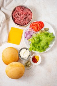 A bowl of ground beef, two cheese slices, a plate of lettuce, tomatoes, pickles, and red onion slices, bowls of mayo, ketchup, mustard, and relish, and two toasted burger buns.