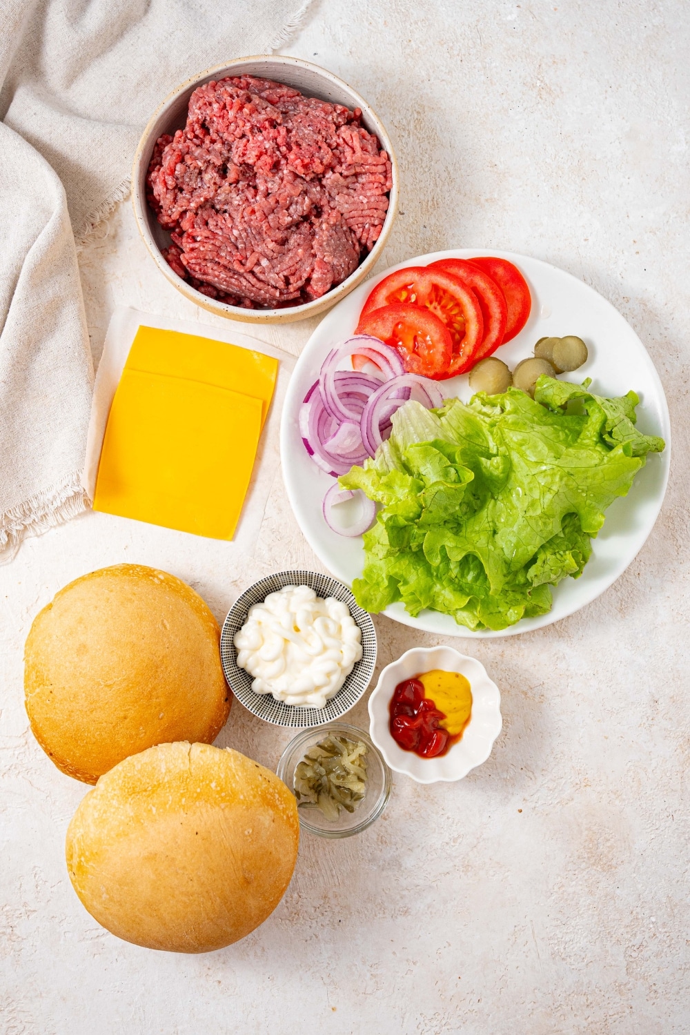 A bowl of ground beef, two cheese slices, a plate of lettuce, tomatoes, pickles, and red onion slices, bowls of mayo, ketchup, mustard, and relish, and two toasted burger buns.