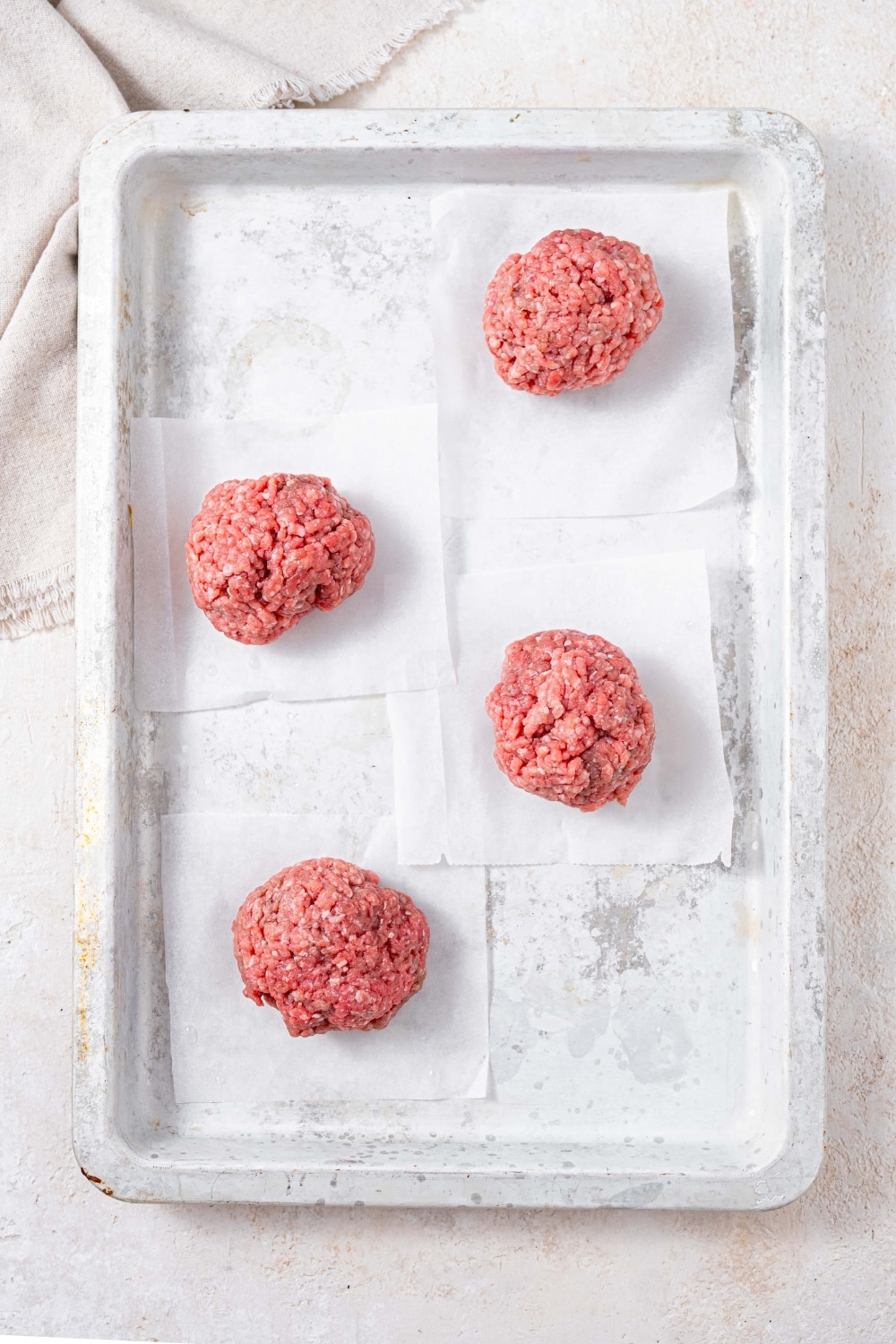 Raw ground beef rolled into balls, arranged on a baking sheet with a piece of parchment paper under each one.