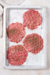 Smashed raw burger patties with seasoning on a baking sheet.