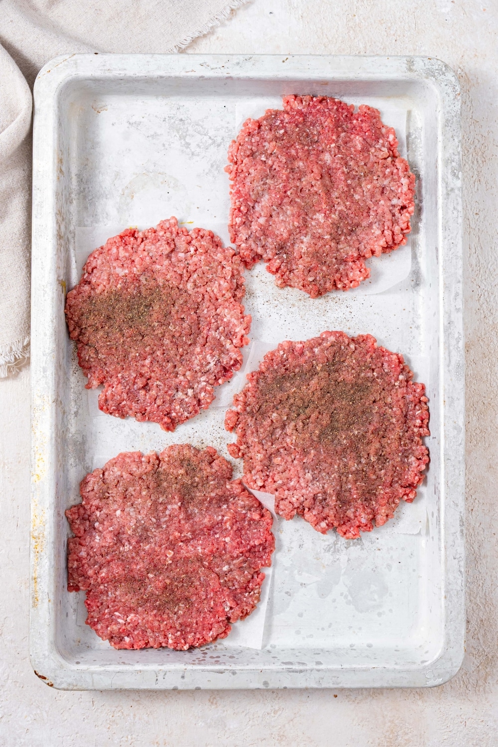 Smashed raw burger patties with seasoning on a baking sheet.
