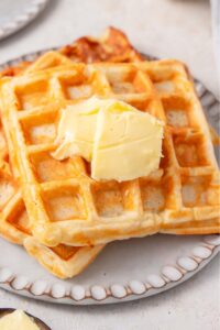 Three cottage cheese waffles on a ceramic plate topped with butter. The plate is on a marble counter.