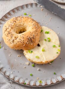 A cottage cheese bagel with everything bagel seasoning and butter spread. The bagel is on a grey ceramic plate garnished with sliced green onion.