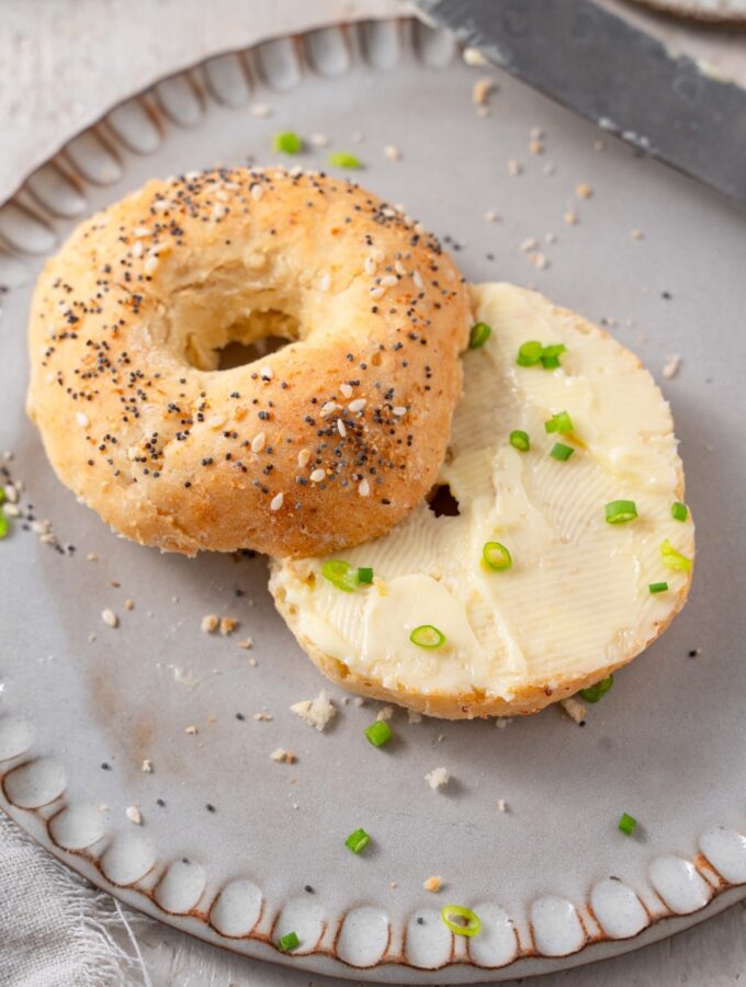 A cottage cheese bagel with everything bagel seasoning and butter spread. The bagel is on a grey ceramic plate garnished with sliced green onion.