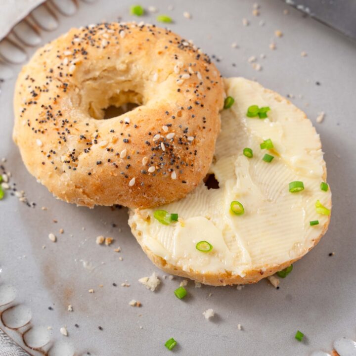 A cottage cheese bagel with everything bagel seasoning and butter spread. The bagel is on a grey ceramic plate garnished with sliced green onion.