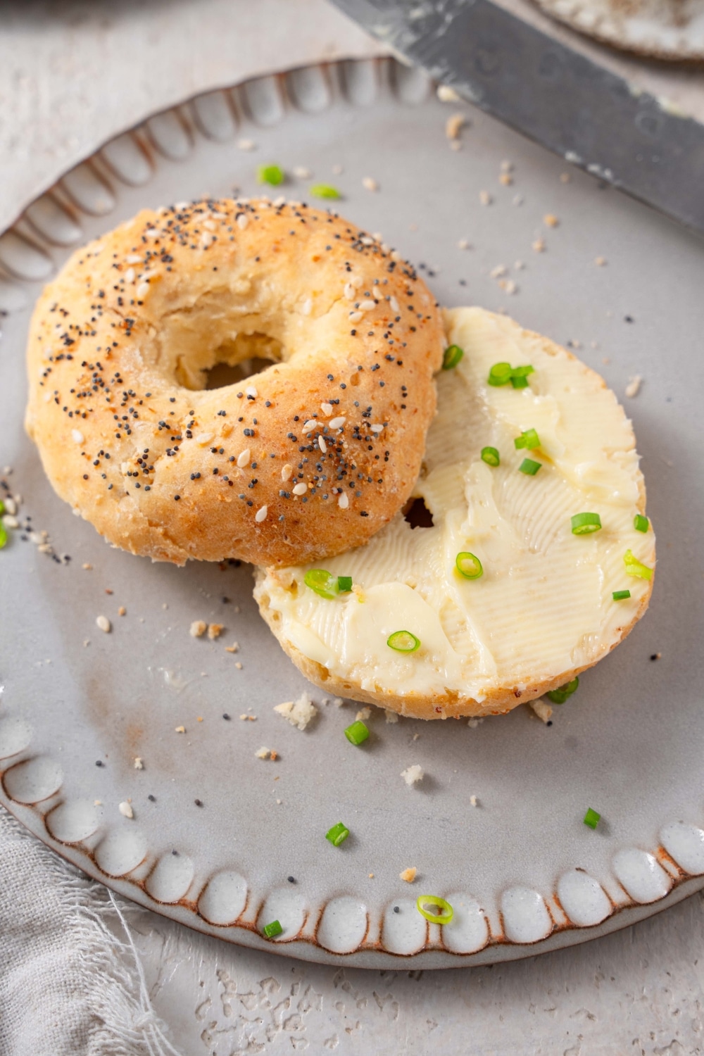A cottage cheese bagel with everything bagel seasoning and butter spread. The bagel is on a grey ceramic plate garnished with sliced green onion.