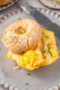 A scrambled egg breakfast sandwich with cottage cheese bagels with everything bagel seasoning. The bagel is on a ceramic plate garnished with sliced green onion and a knife on the side.