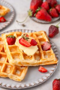 Two cottage cheese waffles on a ceramic plate topped with butter and sliced strawberries. There is a plate of strawberries in the background.