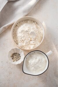 An overhead shot of three bowls of various shapes and sizes containing the ingredients to make cottage cheese bagels including flour, cottage cheese, and everything bagel seasoning.